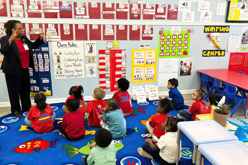 A teacher engages with a group of seated children in a classroom, surrounded by educational posters.