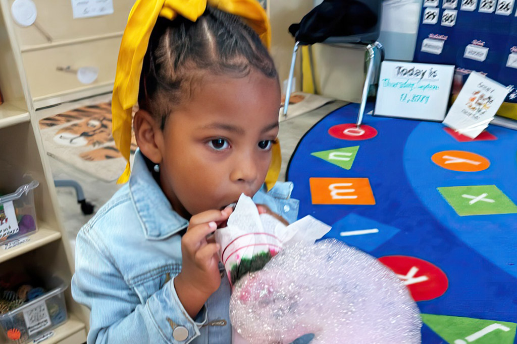 A young girl in a denim jacket and gold colored hair ribbon participates in a classroom science experiment creating sudsy bubbles.