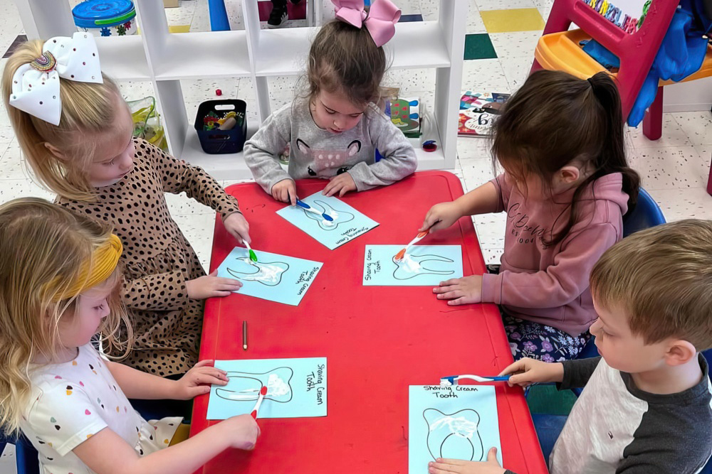 Children gather around a red table, painting tooth designs on blue paper, enjoying a fun art activity together.