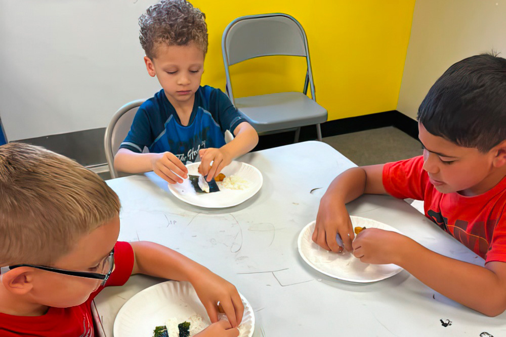 Three boys focus on assembling sushi rolls with seaweed and ingredients on paper plates during a hands-on activity.