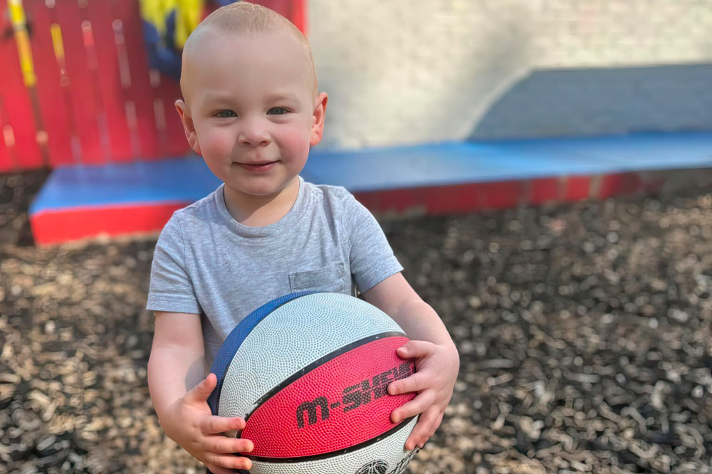 A young child happily poses with a basketball, radiating joy in an outdoor play area.