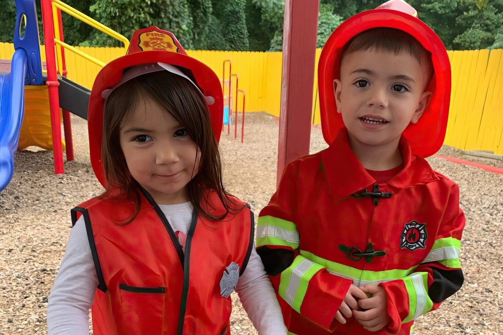 Two children dressed as firefighters smile proudly, wearing bright red hats and jackets in a playground setting.