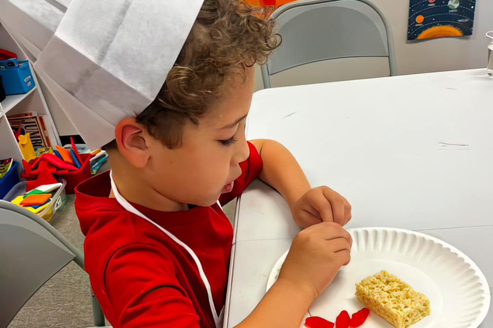 A little boy in a chef hat focuses on decorating a rice treat with red petals at a classroom table.