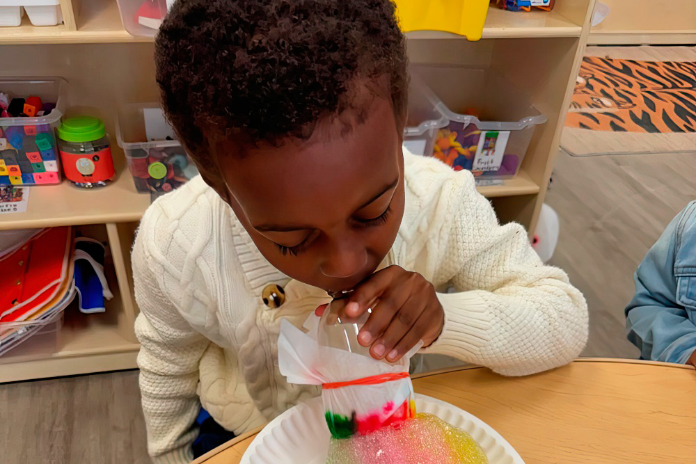 A young boy intently making a bubble from a cup, enjoying a playful and vibrant classroom environment.