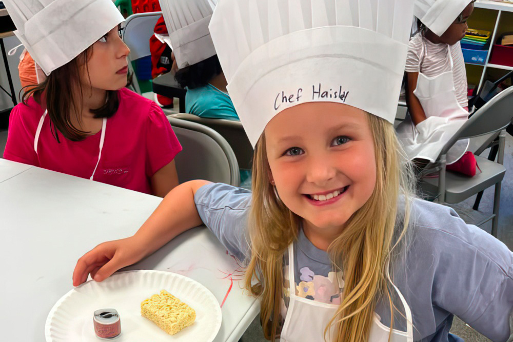 A girl in a chef's hat beams with joy while presenting her culinary creation.