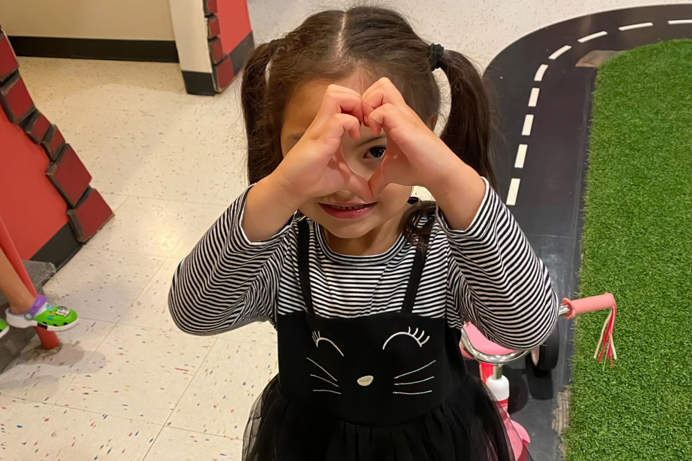 A girl playfully makes a heart shape with her hands, smiling brightly at the camera while wearing a cute dress.