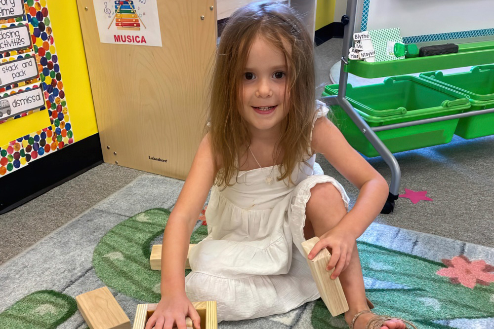 A girl in a white dress is sitting on the floor, smiling while playing with wooden blocks in a colorful classroom.