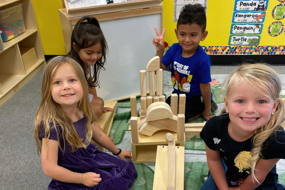 A group of four children smiles while playing with wooden blocks, proudly showcasing their creative structure on the floor.