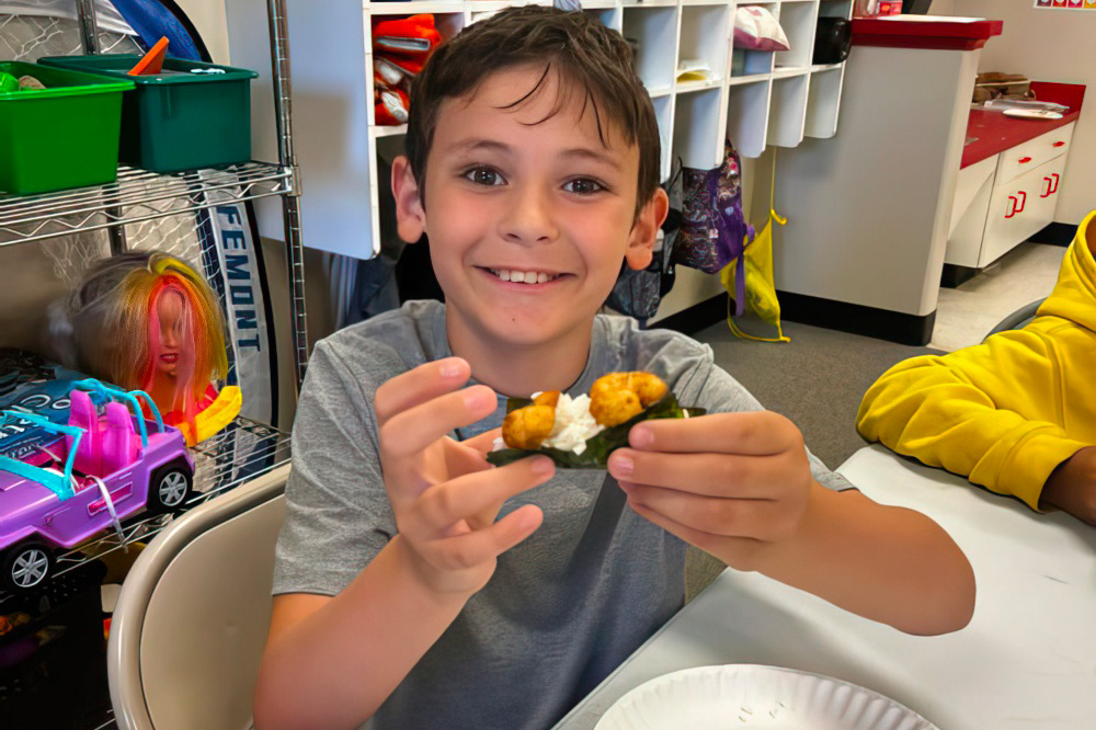 A boy proudly holds up a food item he's made, displaying a sense of accomplishment and enjoyment.