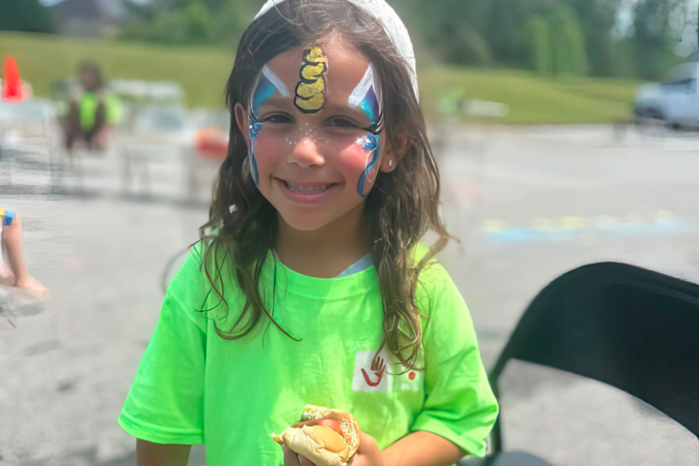 A girl with colorful face paint beams happily, holding a treat.