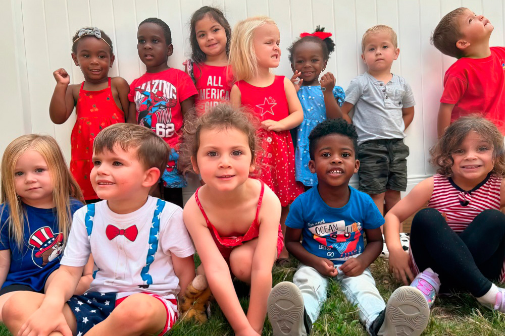 A lively group of children poses together, with smiles and playful expressions against a white fence backdrop.