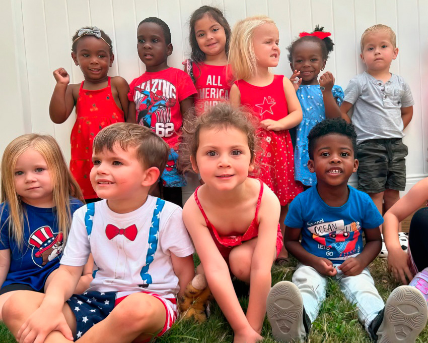 A lively group of children poses together, with smiles and playful expressions against a white fence backdrop.