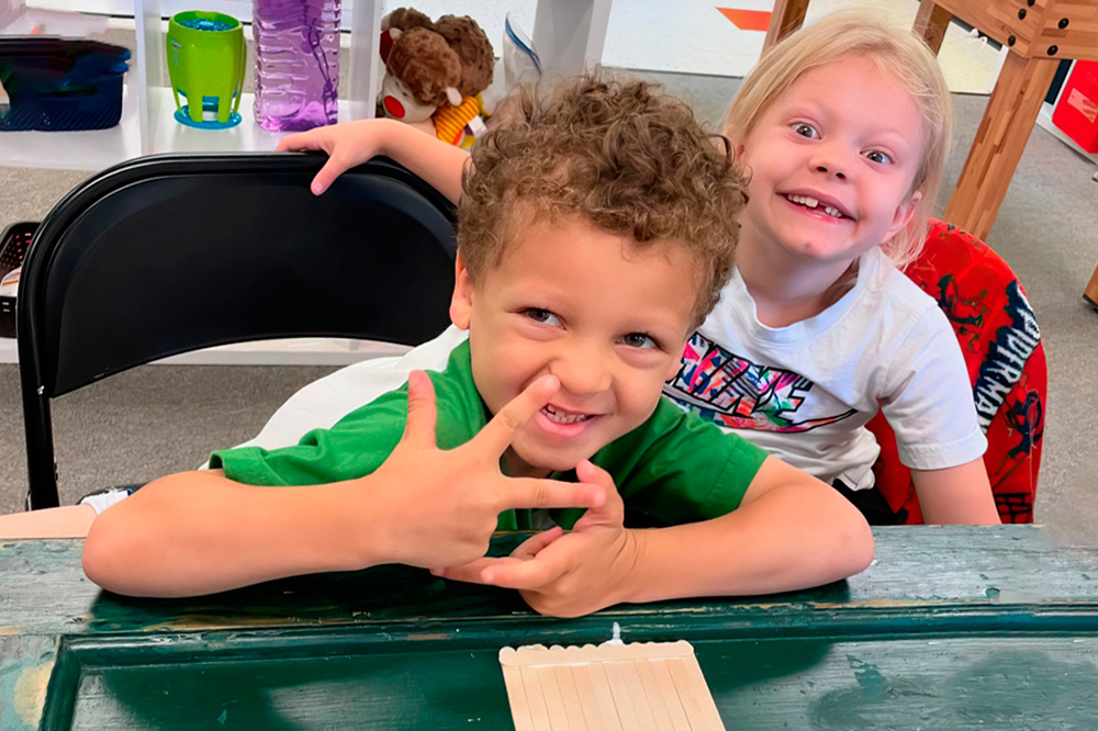 Two young boys sitting together at a table use craft sticks to build a work of art while displaying their playful silliness.