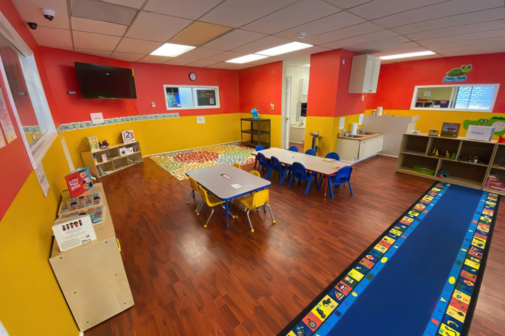 A classroom setting with red and yellow walls includes tables and chairs, designed for a welcoming learning environment.